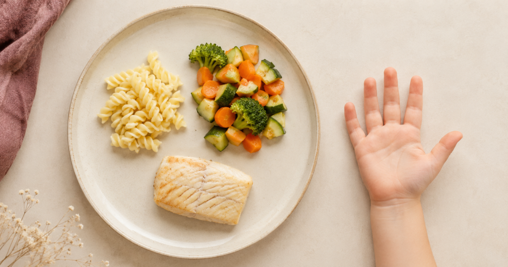 Teller von oben mit einer Portion Nudeln, gedämpftem Gemüse und einem Stück Fisch, daneben eine geöffnete Kinderhand als Größenvergleich
